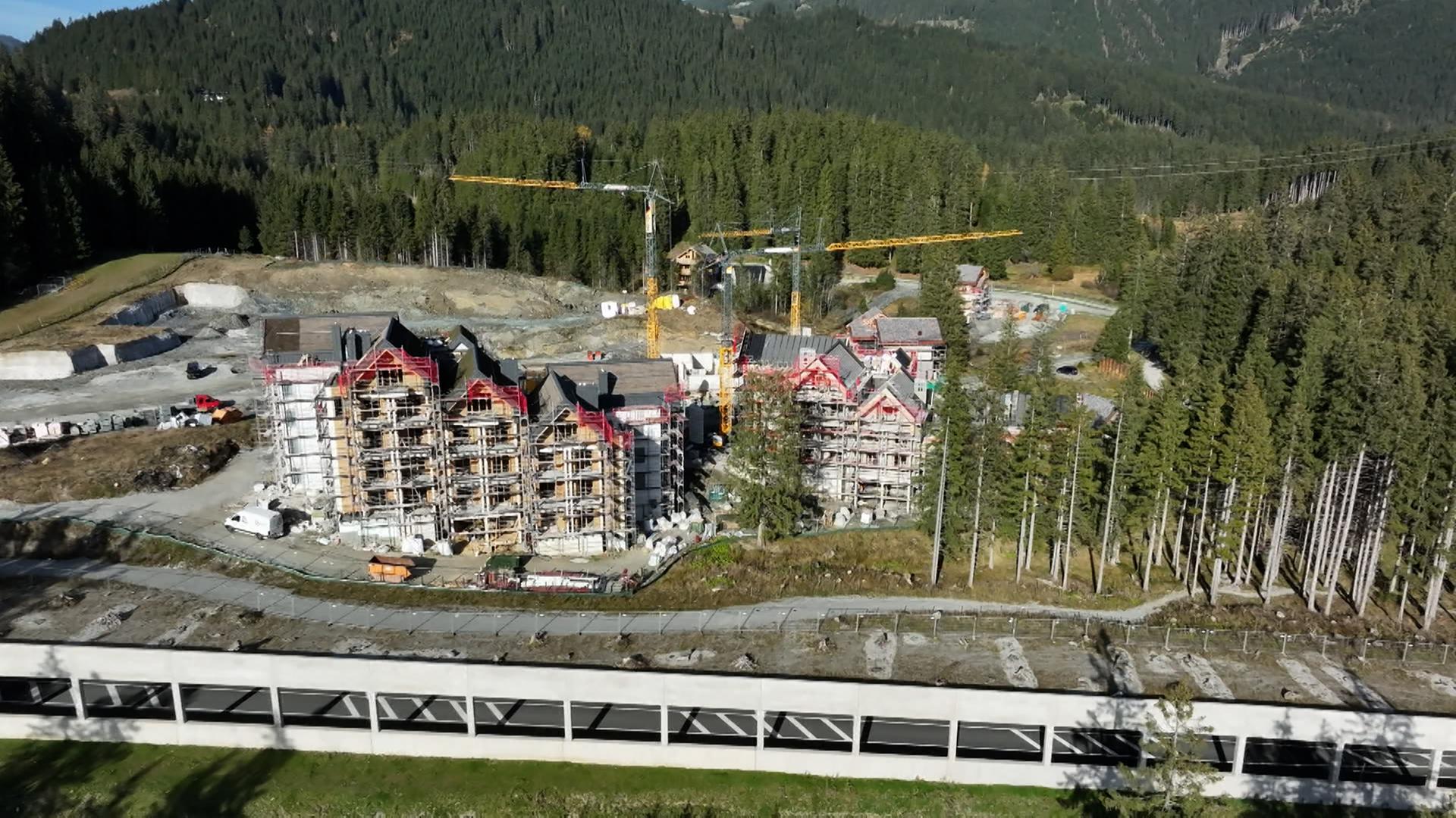 Das Bild zeigt eine Baustelle in den Alpen, konkret am Pass Thurn. Im Vordergrund sind unvollendete Gebäude eines Luxusresorts zu sehen, die von Gerüsten umgeben sind. Einige Teile der Fassaden sind bereits sichtbar, während andere noch Rohbauzustand haben. In der Umgebung sind Kräne aufgestellt, die für den Bau verwendet werden.   Der Untergrund ist teilweise unebenen und es gibt Stellen mit Schotter. Die Landschaft im Hintergrund ist von dichten Wäldern geprägt, die sich bis zu den Berggipfeln erstrecken. Auf der linken Seite des Bildes ist eine Straße sichtbar, die entlang der Baustelle verläuft. Diese Umgebung vermittelt den Eindruck eines stillgelegten, unfertigen Projekts, das sich gegen die natürliche Kulisse der Alpen abhebt.