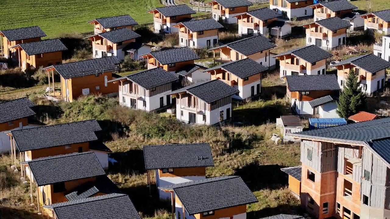 Das Bild zeigt eine große Gruppe von unvollendeten Chalets in einer alpinen Landschaft. Die Chalets haben orangefarbene und weiße Wände sowie schwarze Dächer. Einige der Gebäude scheinen fertiggestellt zu sein, während andere noch im Rohbau sind und keine Fenster oder Türen haben. Die Umgebung ist grün und bewachsen mit Gras und Sträuchern, was auf eine ländliche Lage hinweist. Es sind mehrere Gebäude in unterschiedlichen Anordnungen zu sehen, und die Szenerie vermittelt ein Gefühl von Verlassenheit, da die Immobilien wohl aufgrund eines Baustopps stehen geblieben sind. Die Aufnahme ist aus der Luft gemacht, was einen Überblick über das gesamte Gelände ermöglicht.