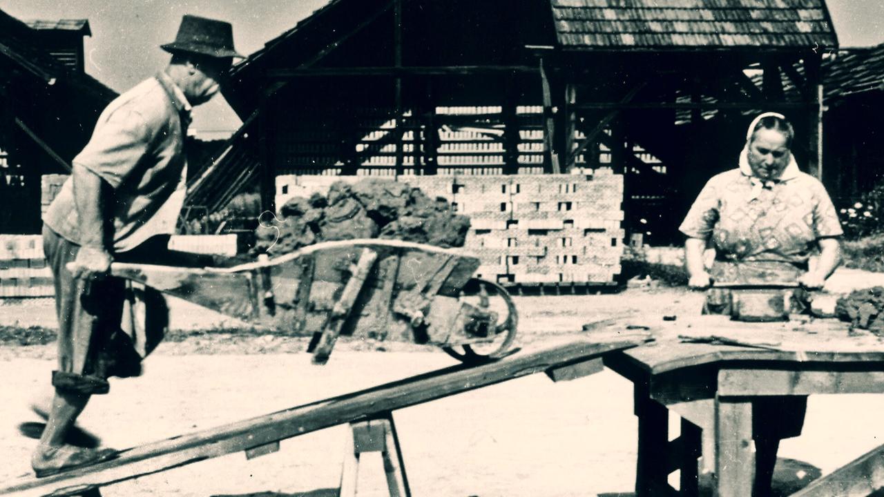 Ein Mann mit Schubkarren und eine Frau an einem Arbeitstisch stehend