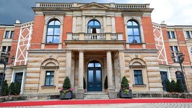 Ein großes zweistöckiges historisches Gebäude aus roten Backsteinen und sandfarbenem Stein mit Säuleneingang und Balkon im oberen Stockwerk.