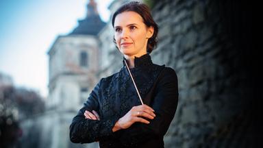 Eine Frau mit kurzen dunklen Haaren und in dunkler Kleidung vor einem alten Gebäude mit Turm