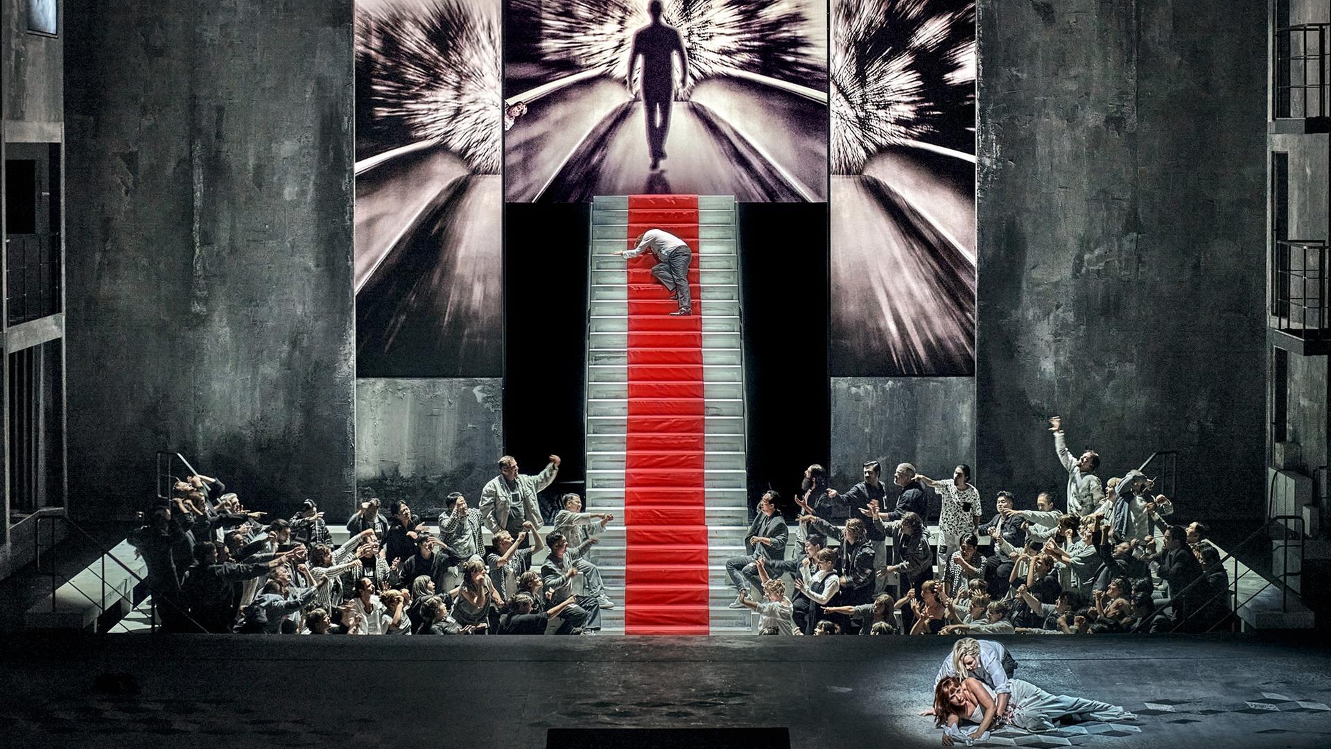 Bühne mit steiler Treppe und zahlreichen Akteuren, die zur Treppe hinweisen, auf der ein Mensch hochkrabbelt