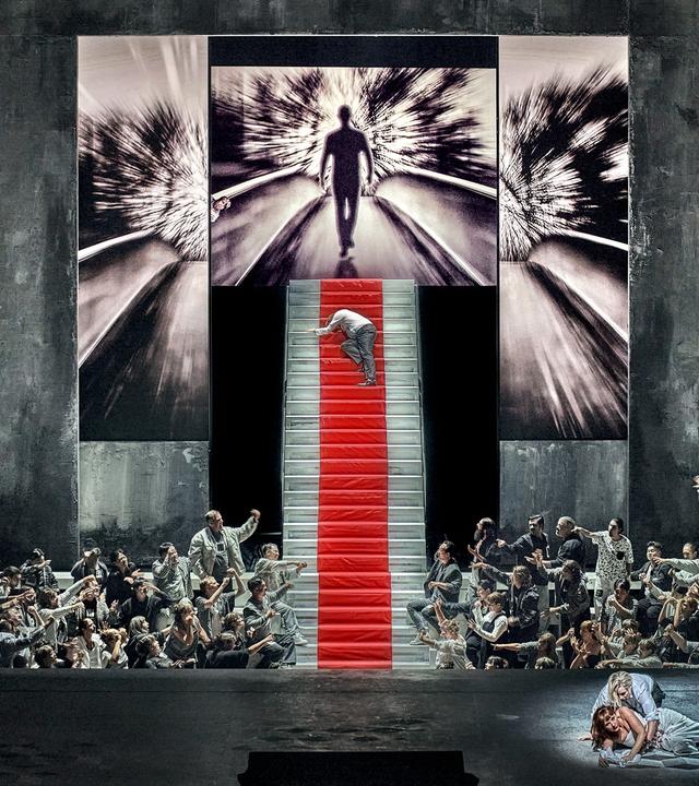 Bühne mit steiler Treppe und zahlreichen Akteuren, die zur Treppe hinweisen, auf der ein Mensch hochkrabbelt