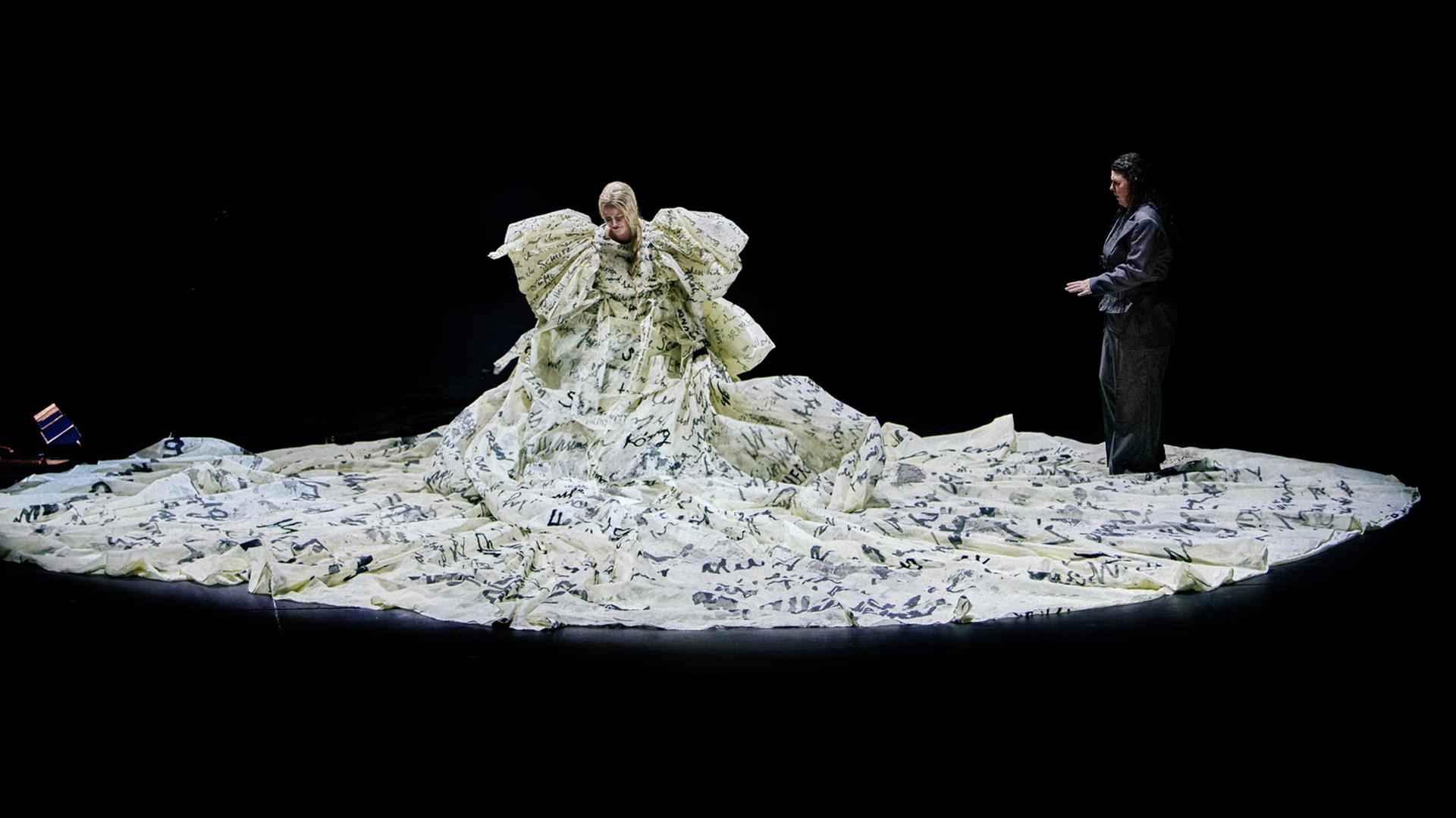 Eine Frau in einem voluminösen hellen Kleid mit Schrift auf einer dunklen Bühne. Neben ihr eine weitere, schwarz gekleidete Frau.