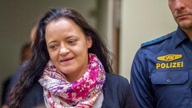 Eine Frau mittleren Alters mit langen Haaren und buntem Schal wird von einem Polizisten begleitet.