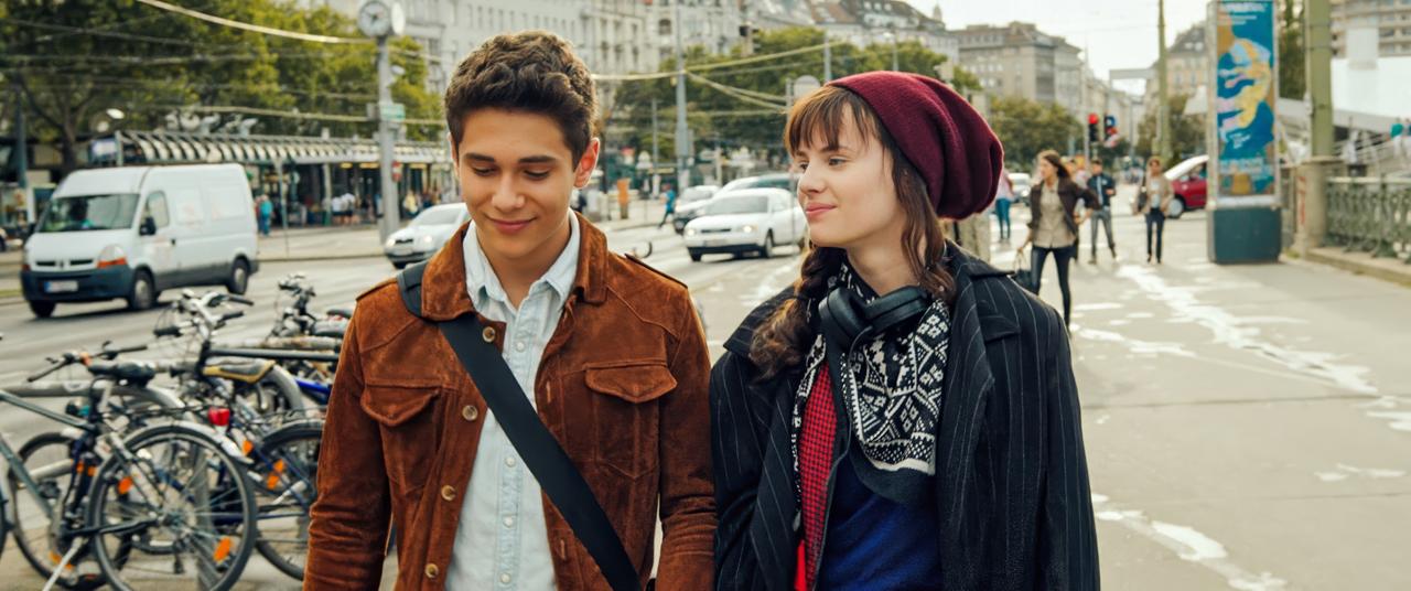 Das Bild zeigt zwei Jugendliche, die gemeinsam auf einer Straße gehen. Es handelt sich um ein urbanes Umfeld mit vielen Fahrrädern im Vordergrund und Häusern im Hintergrund. Der Junge trägt eine braune Wildlederjacke über einem hellen Hemd und hat leicht gewellte, dunkelbraune Haare. Er hat einen freundlichen Gesichtsausdruck und schaut leicht nach unten.   Das Mädchen hat lange, lockige Haare und trägt eine dunkelrote Mütze. Sie hat Kopfhörer um den Hals und einen schwarzen Mantel, der offen getragen wird. Ihre Gesichtszüge sind sanft, und sie lächelt leicht, während sie den Jungen ansieht. Im Hintergrund sind weitere Passanten sowie Fahrzeuge zu sehen, die in beiden Richtungen fahren. Einige Bäume und Straßenschilder sind ebenfalls sichtbar. Insgesamt vermittelt das Bild eine freundliche, jugendliche Stimmung in einer städtischen Landschaft.