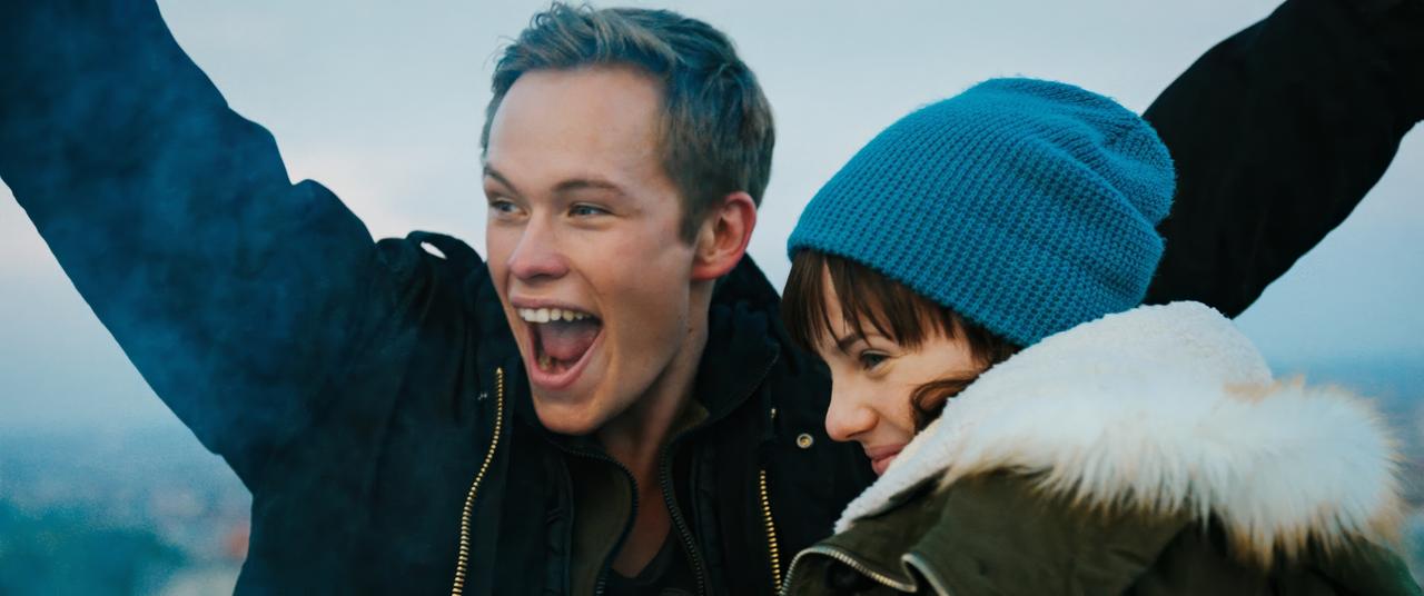 Das Bild zeigt zwei junge Menschen, einen Mann und eine Frau, die fröhlich miteinander interagieren. Der Mann hat kurze, hellblonde Haare und trägt eine dunkle Jacke. Er hebt die Arme in die Luft und lächelt breit, wodurch er Freude und Begeisterung ausstrahlt.   Die Frau hat schulterlange, braune Haare und trägt eine blaue Mütze sowie eine Jacke mit einem pelzbesetzten Kragen. Sie lächelt ebenfalls, wirkt jedoch etwas zurückhaltender in ihrem Ausdruck. Der Hintergrund zeigt einen hellen, wolkigen Himmel, der auf eine tiefere Perspektive hindeutet, möglicherweise von einem erhöhten Standort.   Die Szene vermittelt ein Gefühl von Aufregung und jugendlicher Unbeschwertheit.