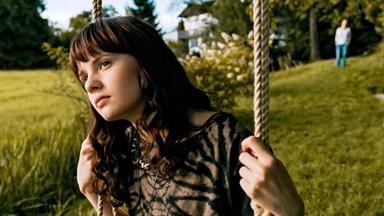 Das Bild zeigt ein junges Mädchen, das auf einer Schaukel sitzt. Sie hat lange, wellige, braune Haare und trägt ein schwarzes, leicht durchscheinendes Oberteil mit einem Muster. Ihr Blick ist nachdenklich und in die Ferne gerichtet. Im Hintergrund ist eine grüne Wiese zu sehen, auf der einige Sträucher und Bäume stehen. Weiter hinten befindet sich ein Gebäude, das möglicherweise ein Wohnhaus ist, und eine Person in heller Kleidung, die in die andere Richtung blickt. Die Szene hat eine ruhige, nachdenkliche Stimmung und wirkt naturnah.