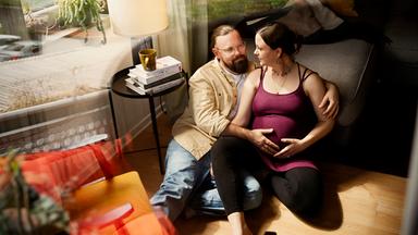 Gitta und Julian sitzen im Wohnzimmer auf dem Boden und schauen sich an. Er hat seine Hand auf ihren Bauch gelegt.