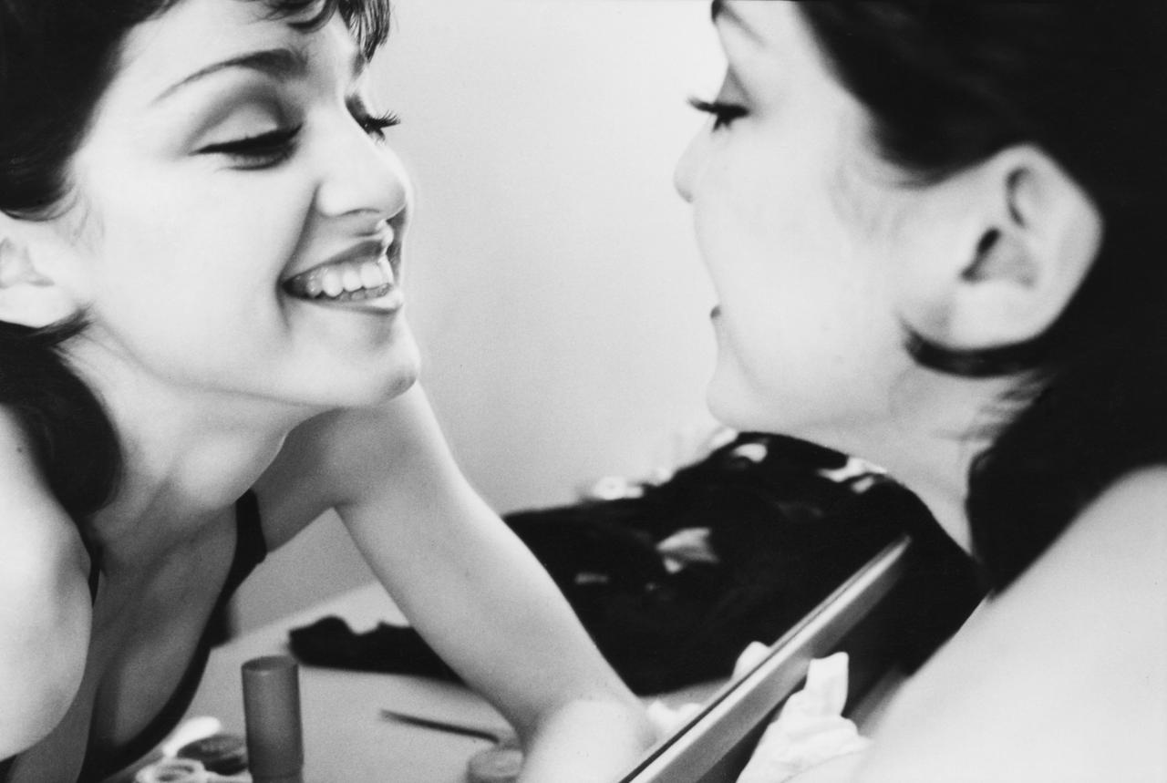 Schwarz-Weiß-Foto: Madonna als junge Frau mit schwarzen Haaren lächelt sich im Spiegel an.