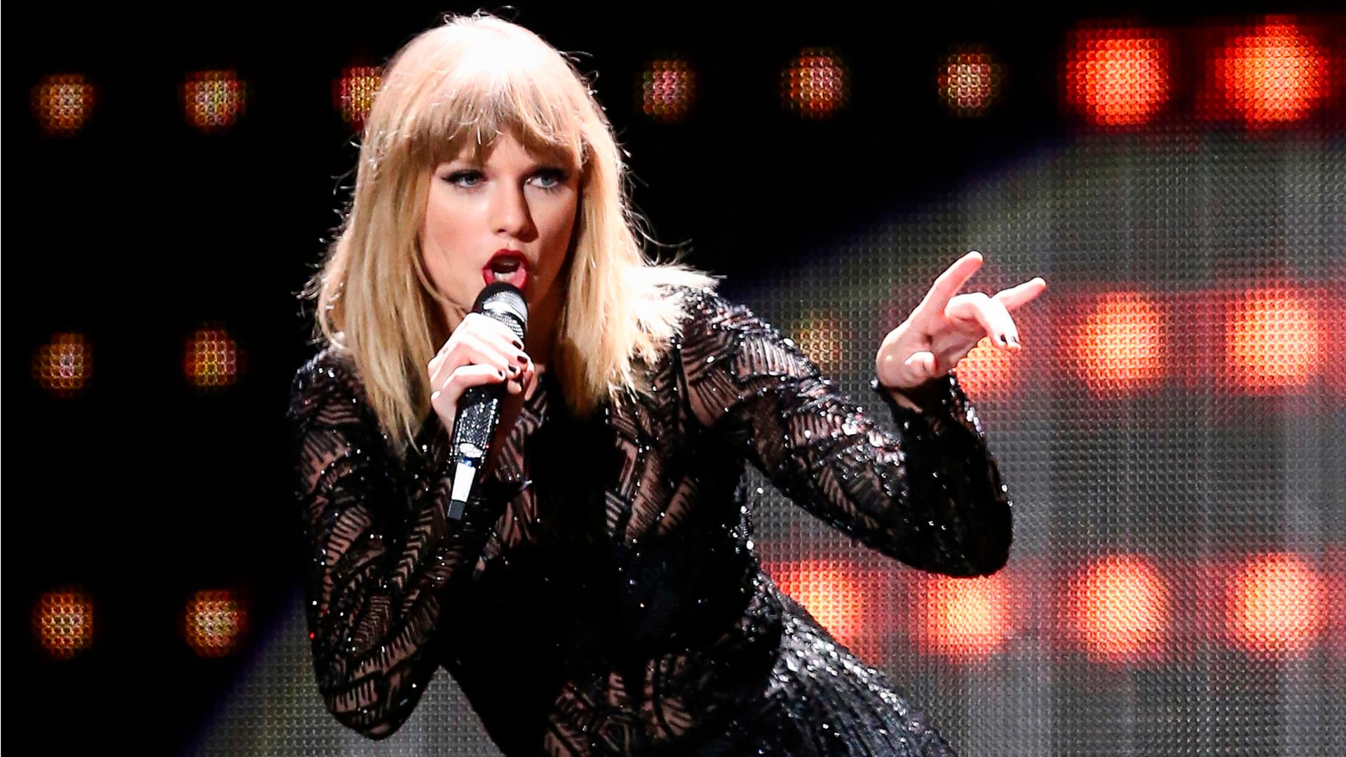 Taylor Swift singt in einem schwarzen Bühnenoutfit mit Mikrofon vor leuchtendem Bühnenhintergrund.