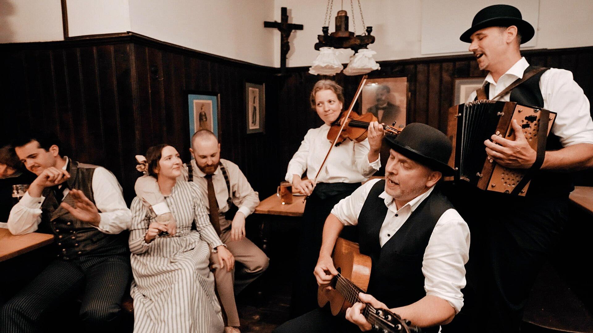Musikanten mit Geige, Gitarre und Ziehharmonika vor  Wirthaus-Publikum