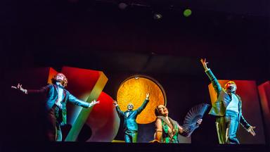 Auf einer dunklen Bühne mit hell angeleuchteten großen Objekten bewegen sich tanzend mehrere Personen.