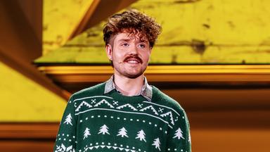 Benedikt Mitmannsgruber steht in der Mitte des Bildes. Der Hintergrund ist gelb beleuchtet und er trägt einen grünen Pullover.  