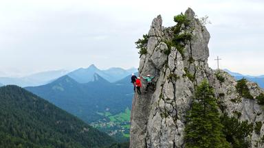 Kletterer an einem senkrechten Felsen. Im Hintergrund stehen bewaldete Berge.