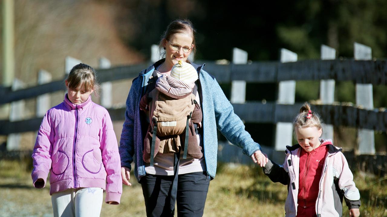 Das Bild zeigt eine Frau mit zwei kleinen Mädchen in einer ländlichen Umgebung. Die Frau trägt ein Baby in einer Tragevorrichtung auf ihrem Bauch und hat eine Brille auf. Sie ist mit einem blauen Oberteil und einem hellen Pullover darunter gekleidet. Die beiden Mädchen sind ebenfalls warm angezogen; eines trägt eine pinke Jacke, während das andere ein rot-weißes Outfit hat.   Im Hintergrund sind eine Holzumzäunung und Weideland zu sehen, das auf eine ländliche Umgebung hindeutet. Die Szene wirkt harmonisch und fröhlich, da die Familie gemeinsam spazieren geht. Es scheint ein sonniger Tag zu sein, was durch das Licht und die Farben des Bildes unterstützt wird.
