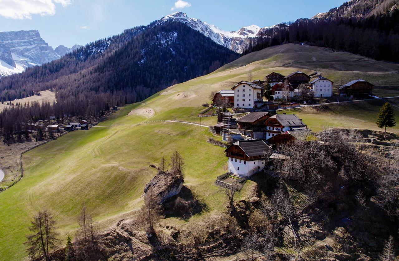 Ein traditionelles Südtiroler Bergdorf mit verstreuten Bauernhäusern auf grünen Hängen vor einer Bergkulisse.