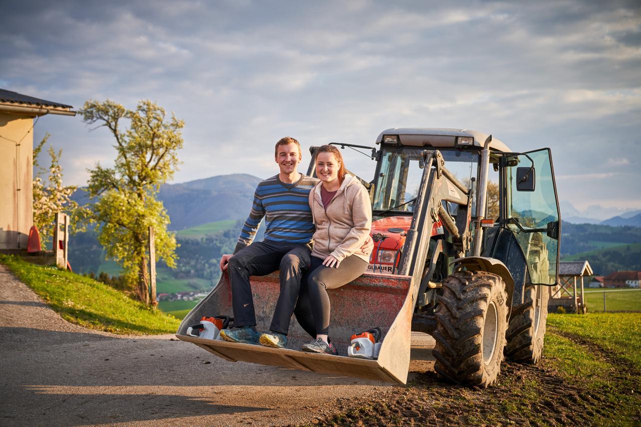 Zwei junge Landwirte, eine Frau und ein Mann, sitzen lächelnd auf der Schaufel eines roten Traktors vor einer malerischen Hügellandschaft.