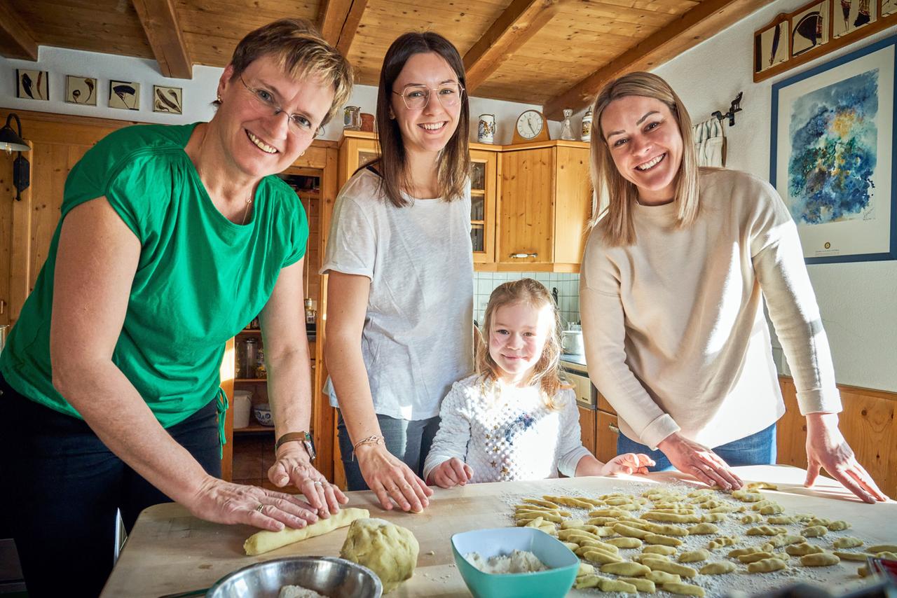 Drei Frauen und ein Mädchen, lächelnd in einer heimeligen Küche stehend, bereiten gemeinsam Teigwaren zu, umgeben von Küchenutensilien und frisch geformten Nudeln.