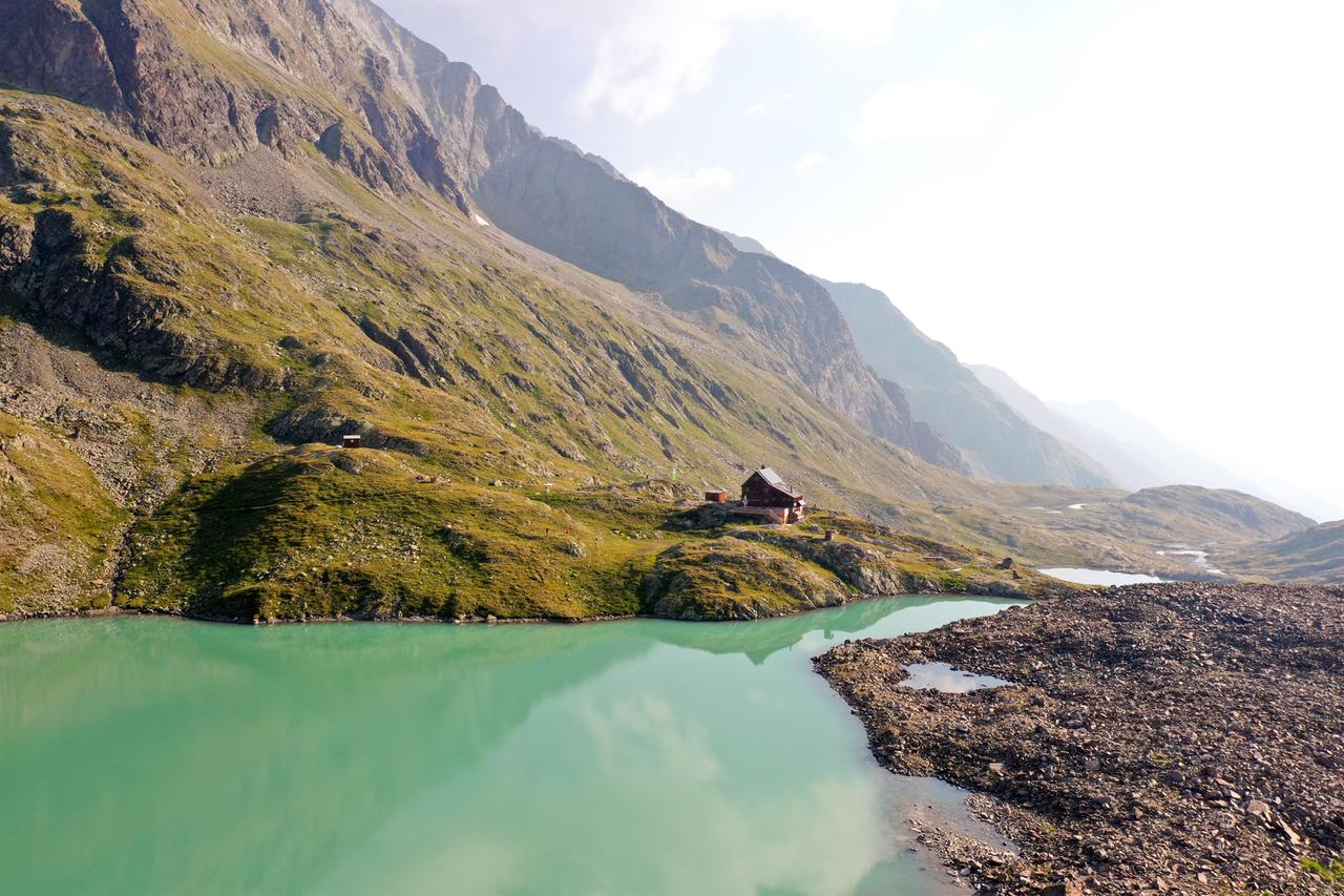 Im Vordergrund ist ein See mit türkisfarbenem Wasser an dem eine kleine Hütte steht. Im Hintergrund sieht man Berge.