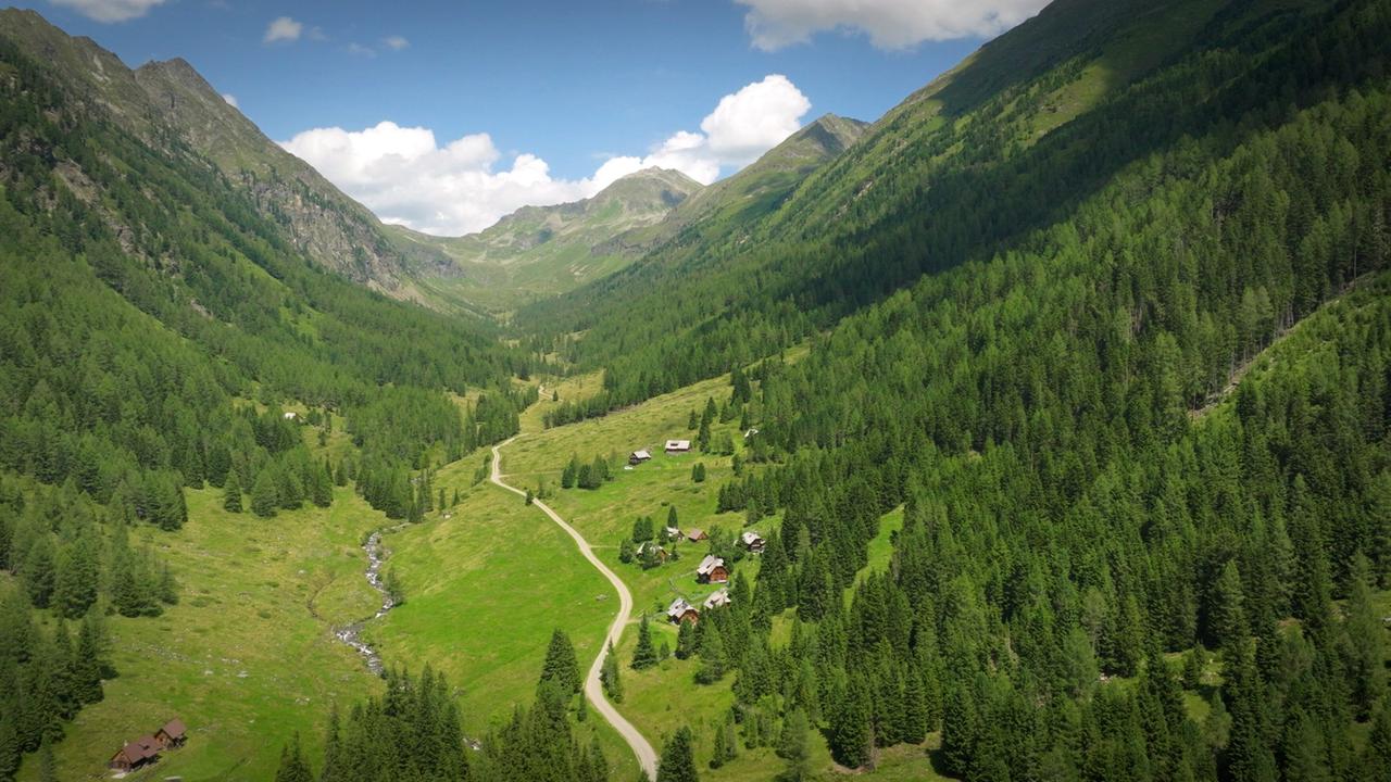 Das Bild zeigt eine malerische Berglandschaft, die von hohen, bewaldeten Bergen umgeben ist. Im Vordergrund erstreckt sich ein sattgrünes Tal mit Wiesen und Niedrigvegetation. Mehrere kleine, traditionelle Hütten und Gebäude sind in der Wiesenfläche verteilt, die in verschiedenen Brauntönen und mit grauen Dächern gestaltet sind. Ein schmaler Weg schlängelt sich durch das Tal und verbindet die Hütten.   Im Hintergrund sind die Berghänge steil ansteigend, bedeckt mit dichter Vegetation und vereinzelten Felsformationen. Die Himmel ist teilweise bewölkt, es sind jedoch auch helle, blaue Abschnitte sichtbar, die für eine freundliche Lichtstimmung sorgen. Der Fluss, der durch das Bild verläuft, ist nur schemenhaft angedeutet, könnte jedoch frisches Wasser führen, das mit dem Landschaftsbild harmoniert.   Insgesamt vermittelt das Bild ein Gefühl von Ruhe und Abgeschiedenheit in der Natur, typisch für eine Bergregion.