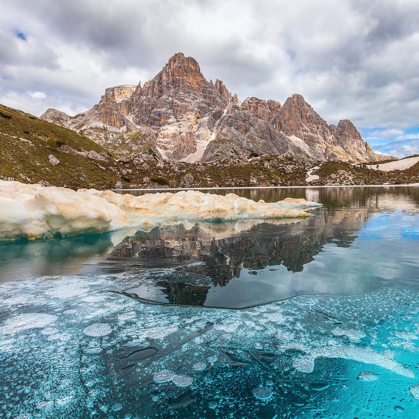 Ein Bergpanorama in der Ferne, im Vordergrund ist ein teilweise gefrorener Bergsee zu sehen.