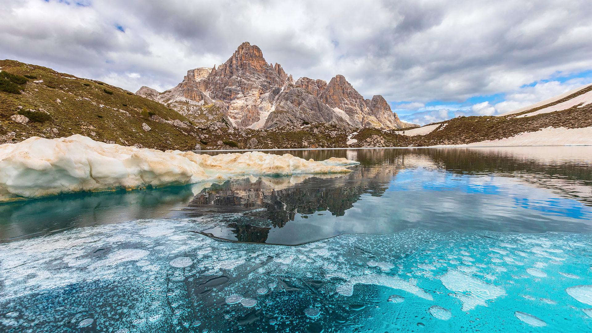 Ein Bergpanorama in der Ferne, im Vordergrund ist ein teilweise gefrorener Bergsee zu sehen.