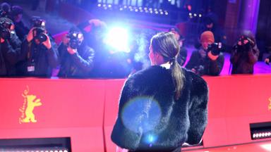 Eine Frau in einer kurzen schwarzen Pelzjacke wendet dem Betrachter den Rücken zu, während sie auf dem Roten Teppich der Berlinale von einer Schaar Fotografen hinter einer Absperrung fotografiert wird.