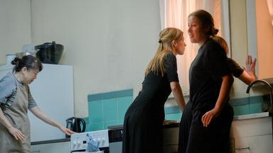 Vier Frauen in einer Küchenkulisse. Links bedient eine Frau in Schürze den Herd. In der Mitte beugt sich eine Frau in schwarzer Kleidung über die Spüle und schaut angespannt nach draußen. Neben ihr steht eine weitere Frau in Schwarz, die sich leicht zur Seite dreht und mit einem Lächeln den Raum betrachtet. Die vierte Frau steht dicht am Fenster und legt eine Hand an die Scheibe, während ihr Blick nach draußen geht. Die Szene wirkt angespannt und geheimnisvoll, geprägt von kühlen Blau- und Grautönen.