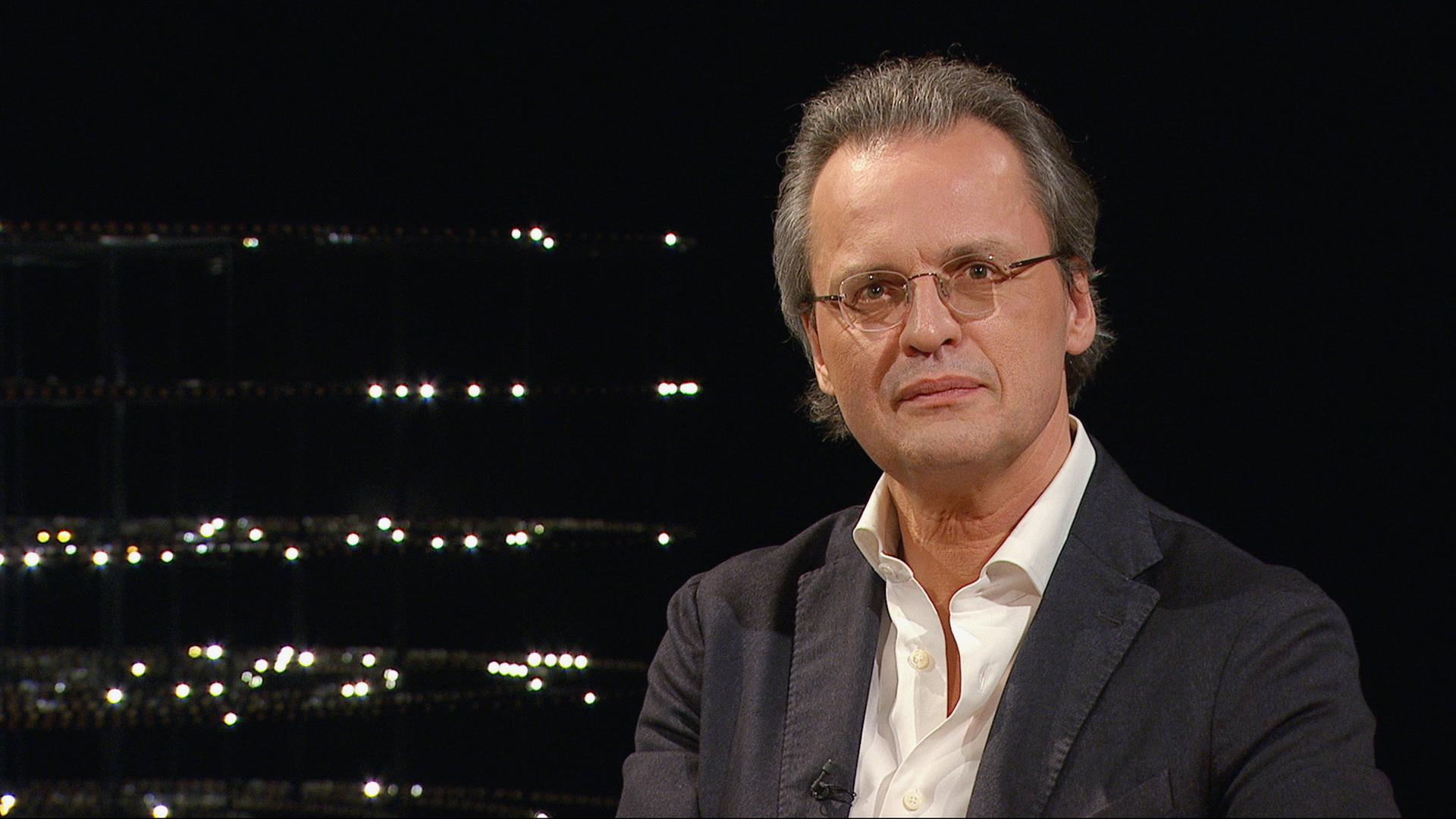 Bernhard Pörksen sitzt in einem dunklen Studio. Er trägt eine Brille, ein dunkles Sakko und ein weißes Hemd. Im Hintergrund sind leuchtende Punkte zu sehen.