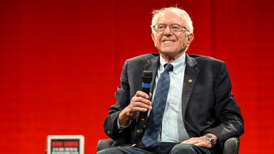 Ein älterer Mann sitzt mit einem Mikro in der Hand in einem Sessel.