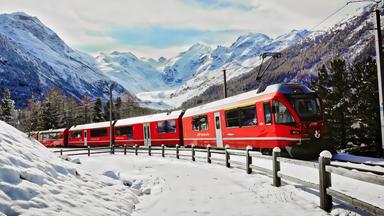 Eine rote Bahn fährt durch eine verschneite Gebirgslandschaft, umgeben von schneebedeckten Bäumen und hohen Bergen.