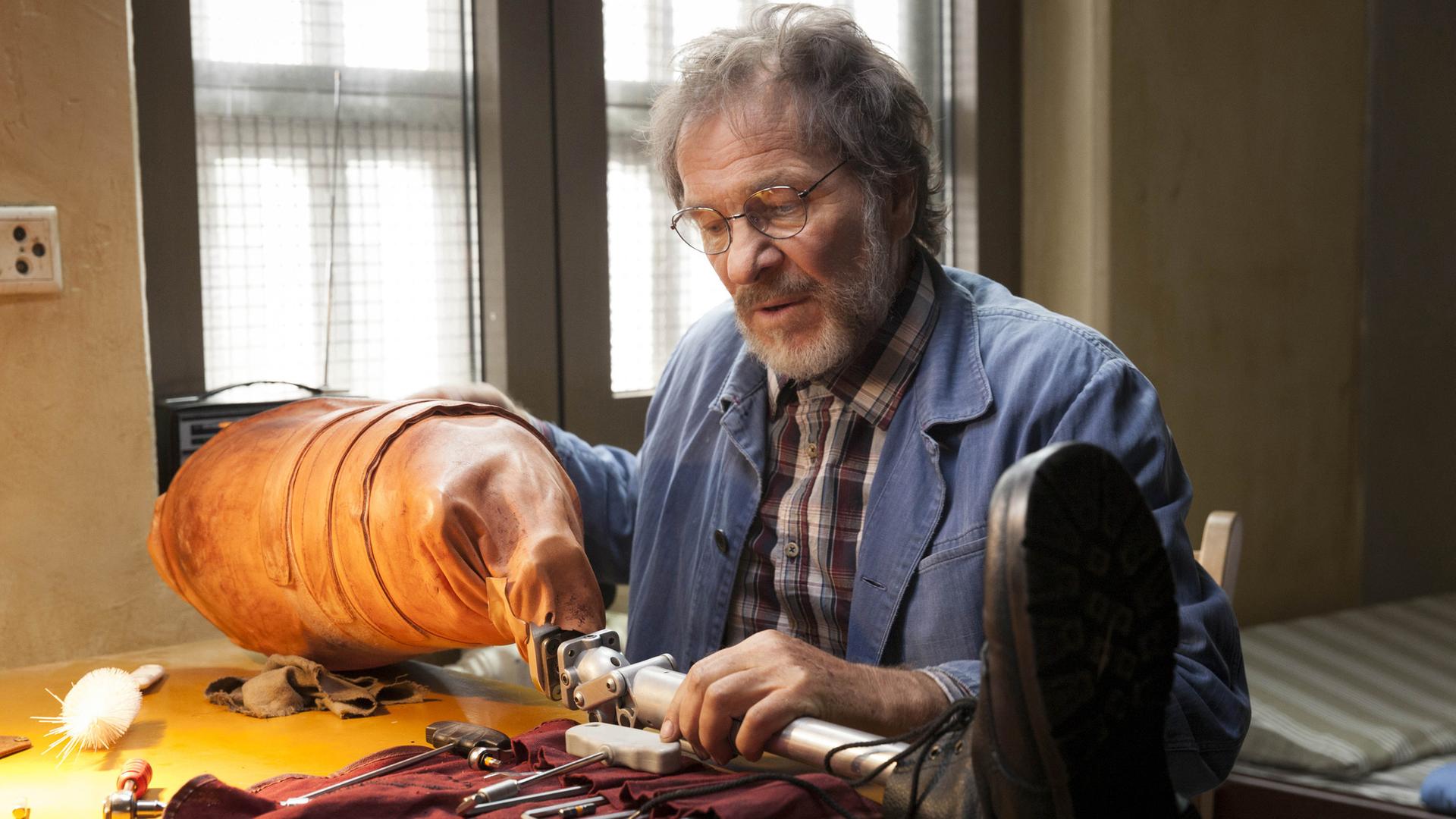 Joseph Komalschek (Götz George) sitzt am Tisch und arbeitet an einer Beinprothese.
