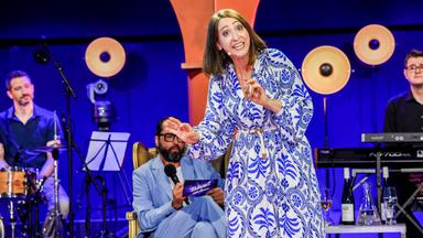Eine dunkehaarige Frau in einem weiß-blau gemusterten Kleid vor einem blauen Hintergrund