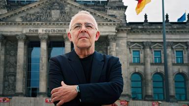 Ein älterer Mann mit Brille und dunkler Kleidung steht mit verschränkten Armen vor dem Reichstagsgebäude in Berlin, im Hintergrund weht eine deutsche Flagge.