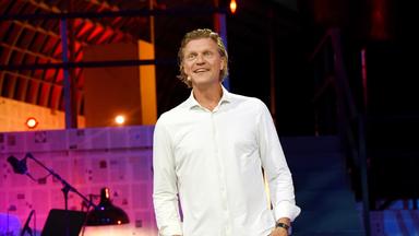Das Bild zeigt einen männlichen Moderator, der auf einer Bühne steht. Er trägt ein weißes Hemd und hat helles, mittellanges Haar. Seine Gesichtsausdruck wirkt freundlich und einladend. Im Hintergrund sind Farbstrahler sichtbar, die die Bühne in warmen Tönen beleuchten. Die Bühne ist einfach gestaltet, mit einem Holzrahmen und einer Reihe von Stufen. Auf der linken Seite des Bildes sind ein Mikrophon-Ständer und eine Lampe zu sehen. Die Gesamtstimmung wirkt festlich und unterhaltsam, passend zu einer kabarettistischen Veranstaltung im Freien.
