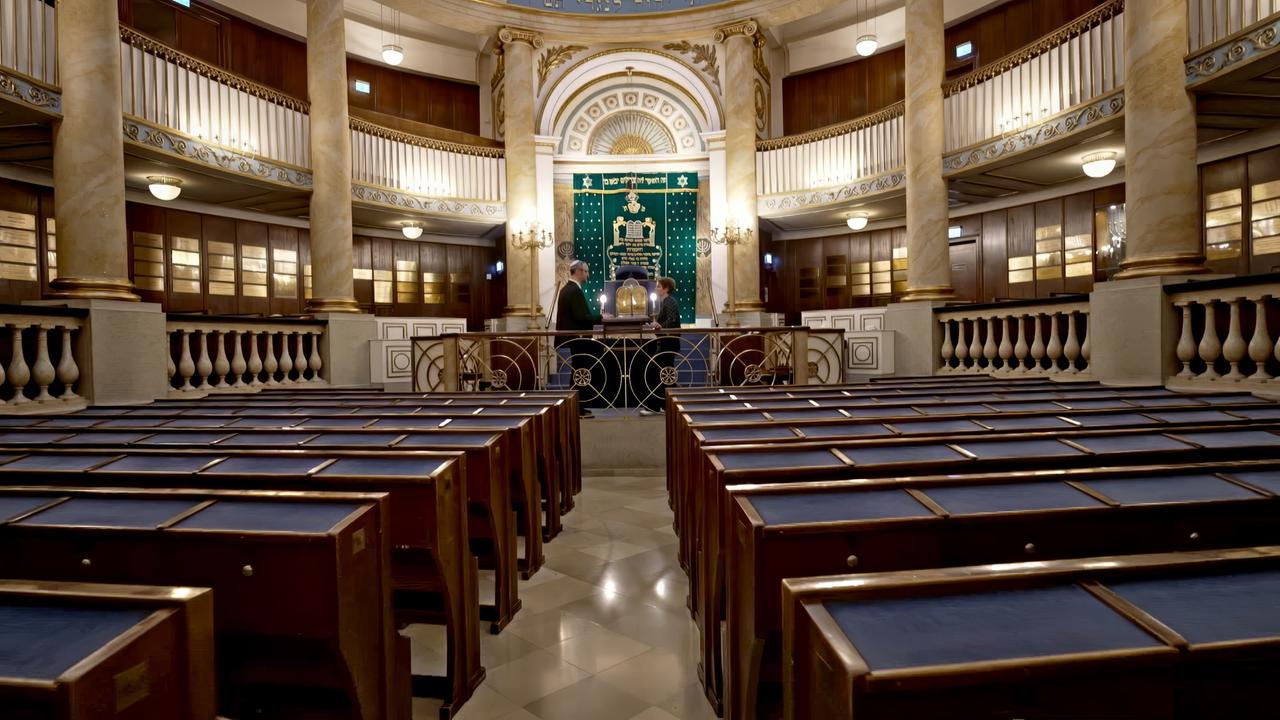 Das Bild zeigt das Innere einer Synagoge in Wien. Der Raum ist symmetrisch angeordnet, mit Holzverkleidung an den Wänden und einer hohen Decke. In der Mitte des Raumes befindet sich eine Bima, die von einem schmiedeeisernen Geländer umgeben ist. Auf der Bima sind zwei Kerzenleuchter zu sehen.  Der Hintergrund wird durch einen großen, grünen Parochet (Vorhang) dominiert, der mit goldenen und blauen Symbolen verziert ist. Auf der rechten und linken Seite sind Sitzbänke aus Holz, die mit blauen Polstern ausgestattet sind, und die Reihen der Bänke ziehen sich bis zur Rückwand des Raumes. Die Wände sind mit goldenen Verzierungen und moderner Beleuchtung ausgestattet.  Im Vordergrund stehen zwei Personen im Gespräch. Das Licht im Raum ist warm und hell, was eine einladende Atmosphäre schafft. In der Synagoge befindet sich nichts, was auf eine spezifische Zeit oder eine bestimmte Handlung hinweist, aber sie vermittelt einen spirituellen und historischen Eindruck.