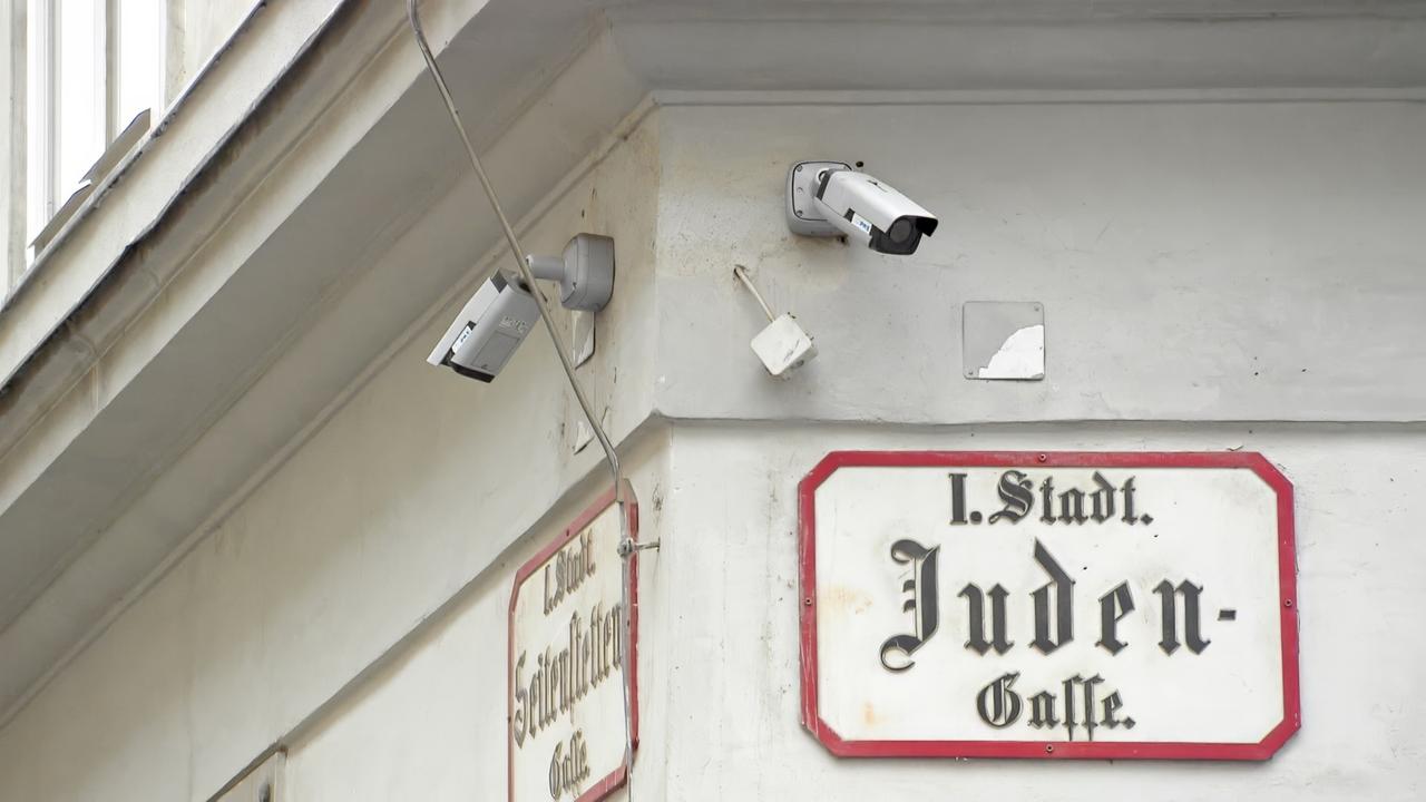 Das Bild zeigt eine Wand mit einem Eingangsschild, das den Namen "Juden-Gasse" in einer klassischen Schriftart enthält. Das Schild ist rot umrandet und gut lesbar. Darüber hängen drei Überwachungskameras, die auf die Gasse gerichtet sind. Die Wände um das Schild sind farblich hell und tragen einige Abnutzungserscheinungen, wie zum Beispiel kleine Flecken oder abgeschlagene Stellen. Im Hintergrund sind auch Fenster zu sehen, die teilweise geöffnet sind. Insgesamt vermittelt das Bild einen Eindruck von Sicherheitsmaßnahmen in einem städtischen Umfeld, das mit einer jüdischen Geschichte verbunden ist.