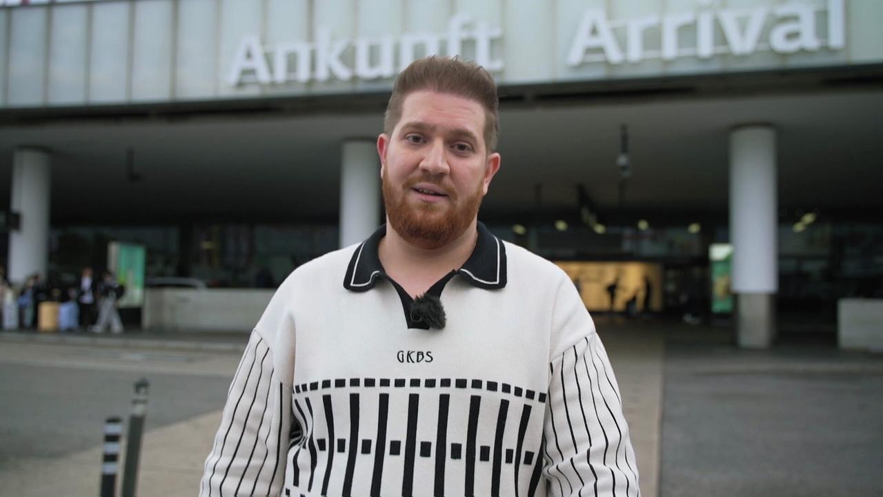 Das Bild zeigt einen Mann, der an einem Flughafen steht. Im Hintergrund ist ein großes Schild mit der Aufschrift „Ankunft“ (auf Deutsch) und „Arrival“ (auf Englisch) zu sehen. Der Mann hat einen roten Bart, trägt ein weißes Sweatshirt mit schwarzem Muster und hat kurzes, welliges Haar. Er steht vor einer modernen Flughafenarchitektur, die durch Säulen und Glasflächen geprägt ist. Im Hintergrund sind einige Personen sichtbar, die sich in einem Wartebereich aufhalten. Der Mann wirkt freundlich und zugewandt. Es ist Tageslicht und die Umgebung scheint geschäftig zu sein, typisch für einen Flughafen.