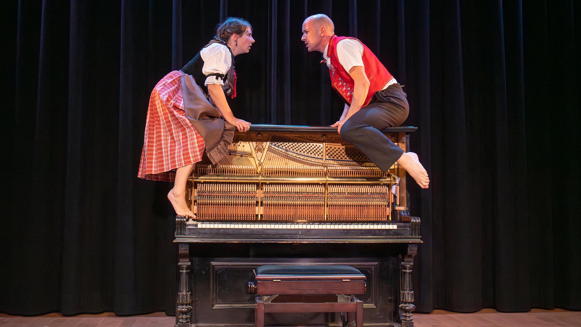 Eine Frau und ein Mann in traditioneller Kleidung sitzen barfuß auf einem Klavier und blicken sich intensiv an. Die Szene spielt auf einer Bühne mit schwarzem Hintergrund.