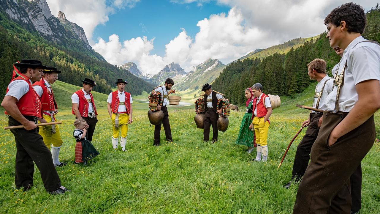 Eine Gruppe von Menschen in Appenzeller Tracht steht auf einer alpinen Wiese. Zwei Männer tragen große verzierte Kuhglocken, umgeben von einer idyllischen Berglandschaft.