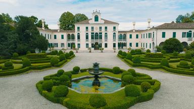 Das Bild zeigt die Villa Tiepolo Passi, eine prächtige Landvilla, umgeben von einem gepflegten Garten. Der zentrale Baukörper der Villa präsentiert sich in hellem Weiß mit eleganten Fenstern, die grüne Fensterläden besitzen. Auf dem Gebäude befindet sich ein zierlicher Dachaufsatz, der der Architektur einen klassischen Touch verleiht.  Vor der Villa erstreckt sich ein großzügiger Garten, der mit kunstvoll geschnittenen Hecken in runden und geschwungenen Formen gestaltet ist. In der Mitte des Gartens befindet sich ein kleiner Teich mit einer dekorativen Brunnenskulptur. Um den Teich sind ebenfalls grüne, üppige Pflanzen angeordnet, die einen harmonischen Gesamteindruck erzeugen.  Im Hintergrund sieht man Bäume, die den Garten umgeben und dem Bild eine natürliche Kulisse verleihen. Der Himmel ist leicht bewölkt, was für eine sanfte Beleuchtung sorgt, und die gesamte Szenerie strahlt eine ruhige, elegante Atmosphäre aus.