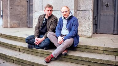 Sebastian Bezzel (links) und Simon Schwarz sitzen auf den Stufen vor dem Eingang des Deutschen Museums