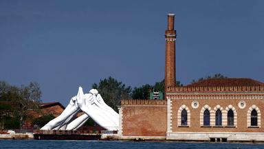 Neben einem alten Ziegelsteingebäude mit Turm am Wasser ist eine große weiße Skulptur zu sehen, die aus Händen besteht, die sie paarweise an der Hand anfassen.