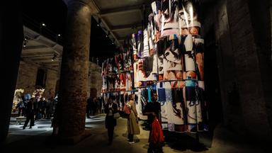 In einem halbdunklen Raum mit hoher Flachdecke laufen Menschen und betrachten ein riesiges Kunstwerk, das aus dreifach hoch gestapelten Bildrollen besteht.