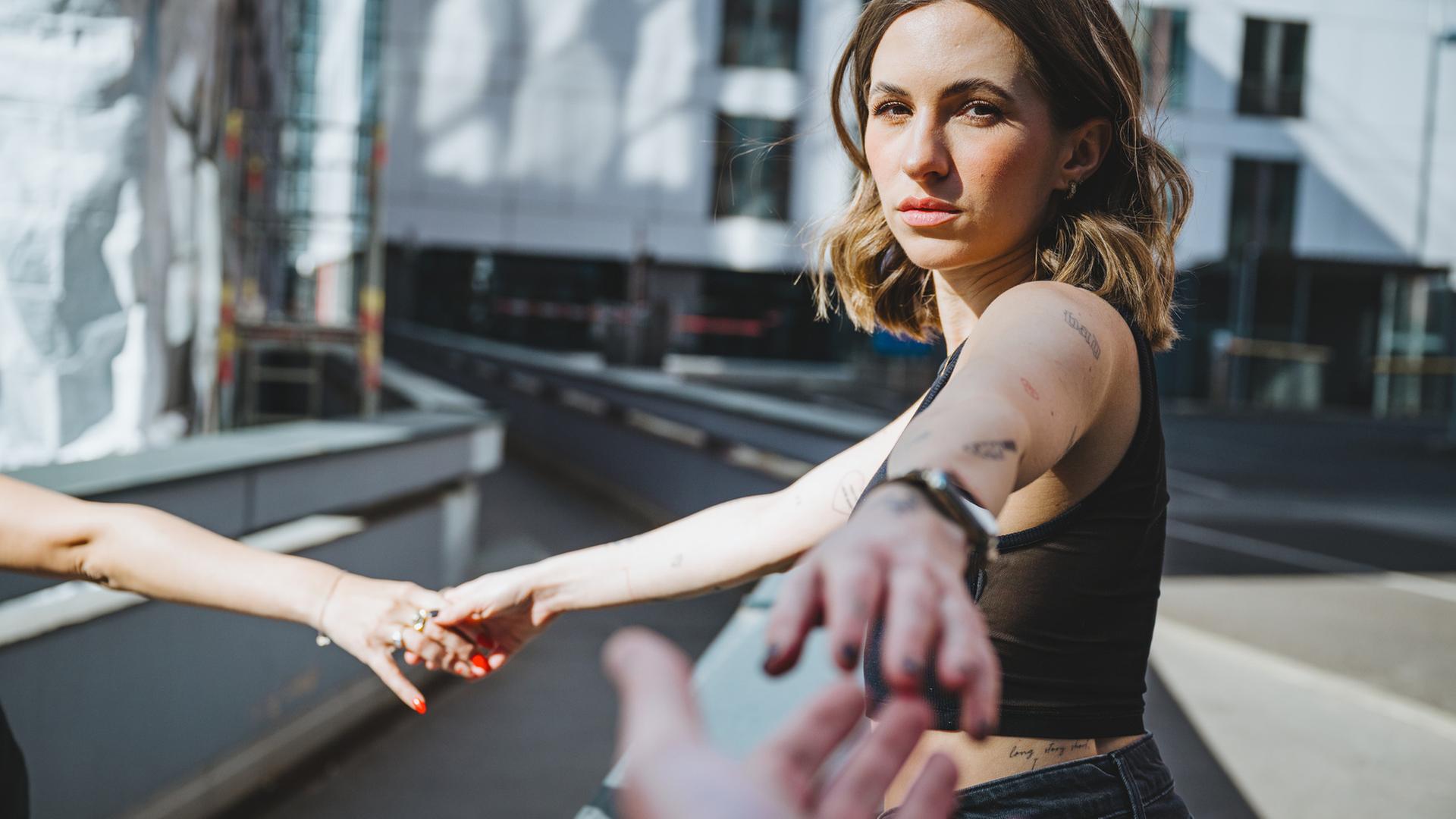 Eine junge Frau entfernt sich mit ausgestrecktem Arm von einer Person außerhalb des Bildrandes und streckt den anderen Arm nach einer dritten Person außerhalb des Bildrandes aus.