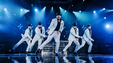 Bühnenstill eines Backstreet Boys Konzertes. Auf der Bühne sind fünf Männer in weißen Anzügen mit schwarzen Hüten zu sehen. 