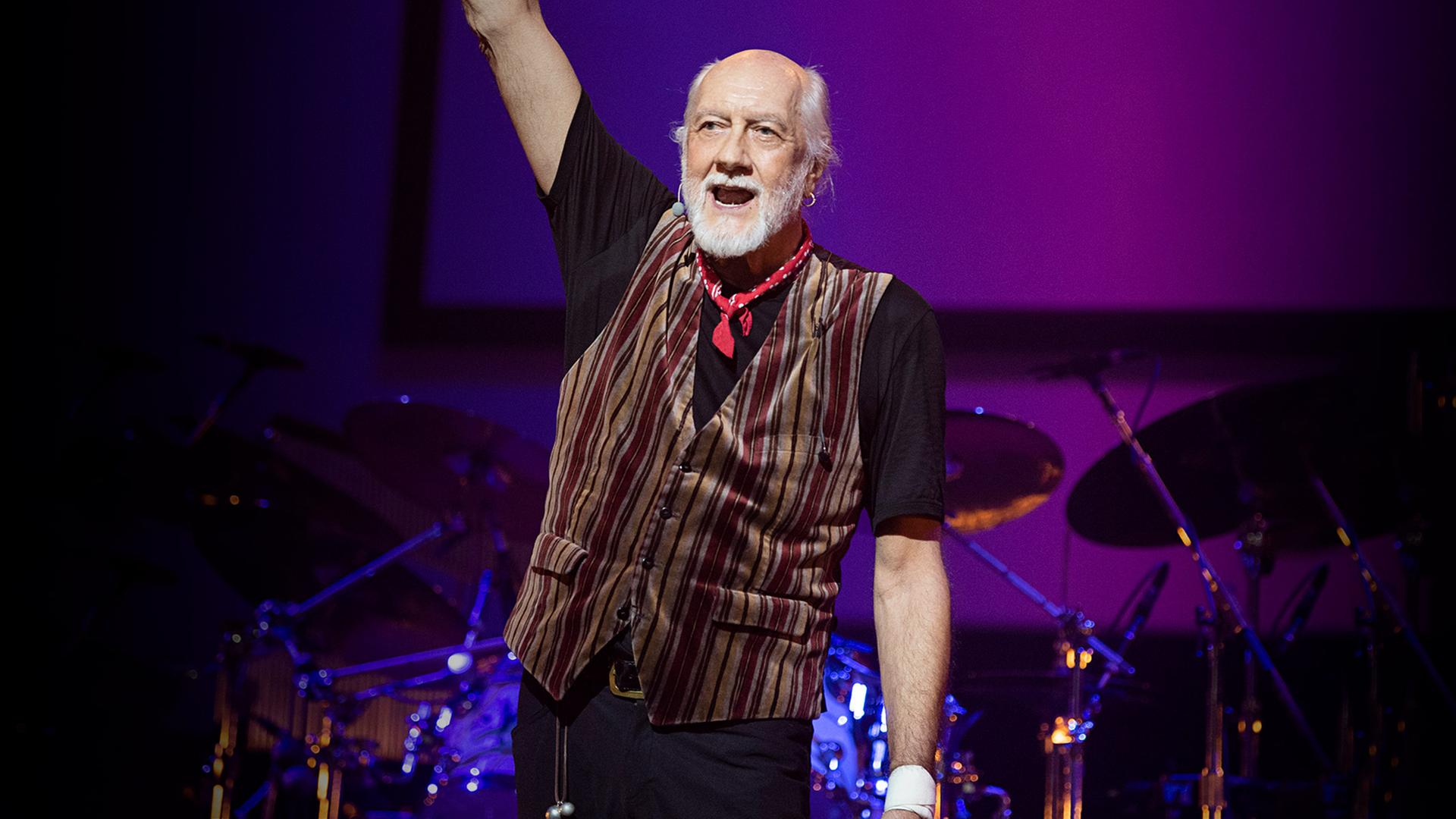 Mick Fleetwood auf der Bühne, er streckt den rechten Arm in die Höhe und hat den Mund geöffnet.