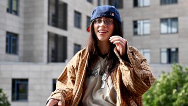 Eine junge Frau in übergroßem Schlabberlook und umgedrehter Baseball-Mütze sitzt vor einem mehrstöckigen Wohnhaus und lacht.