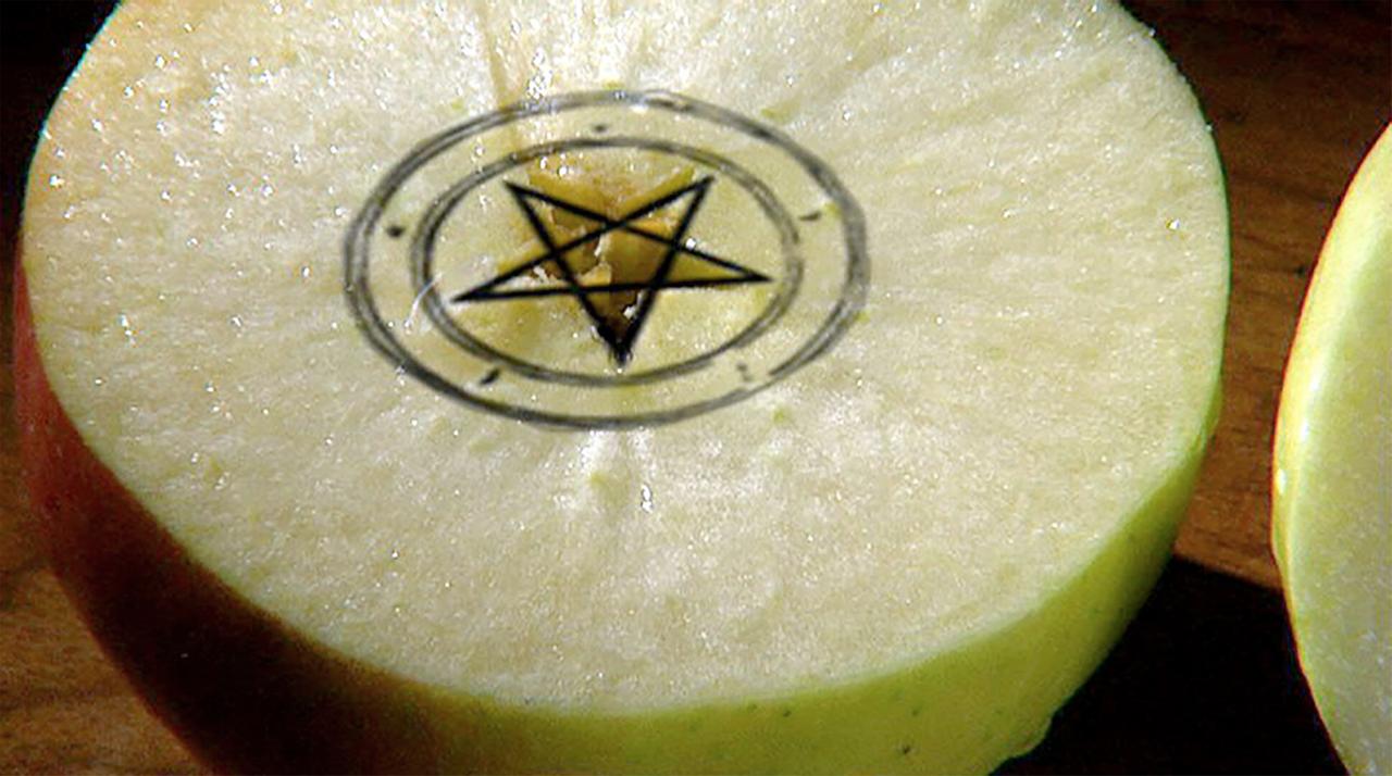 Das Bild zeigt einen halbierten Apfel. In dem Apfel ist ein Pentagramm und ein Kreis abgebildet. Der Apfel hat eine grünlich-weiße Farbe und eine rote Schale.